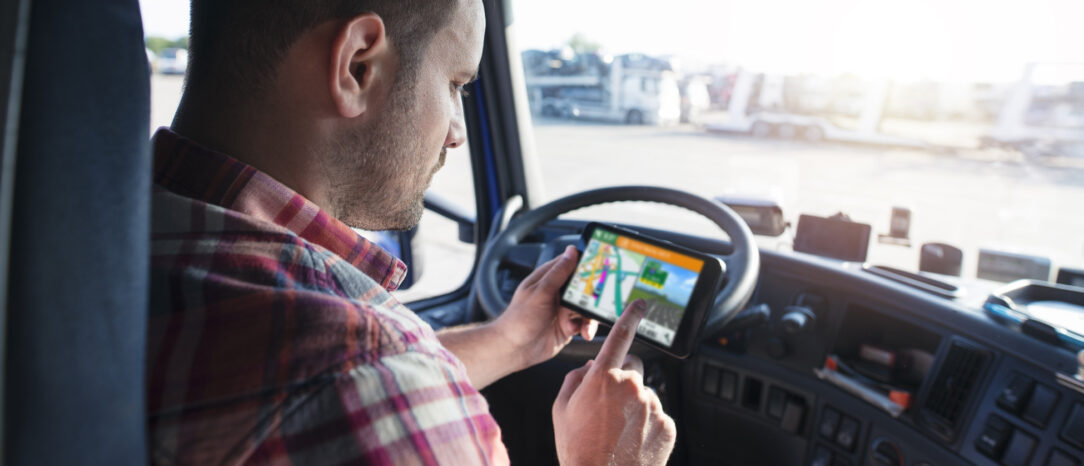 Male truck driver sitting in the cab of a parked truck, checking the GPS on his phone.