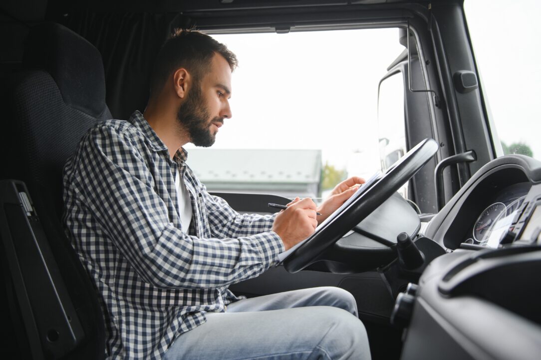 Man in a truck cab shown in profile, wearing a checkered shirt, writing on a clipboard at the driver's seat