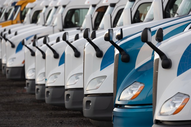 Row of white and blue semi-trucks lined up in a parking lot, facing forward with their extended side mirrors visible.