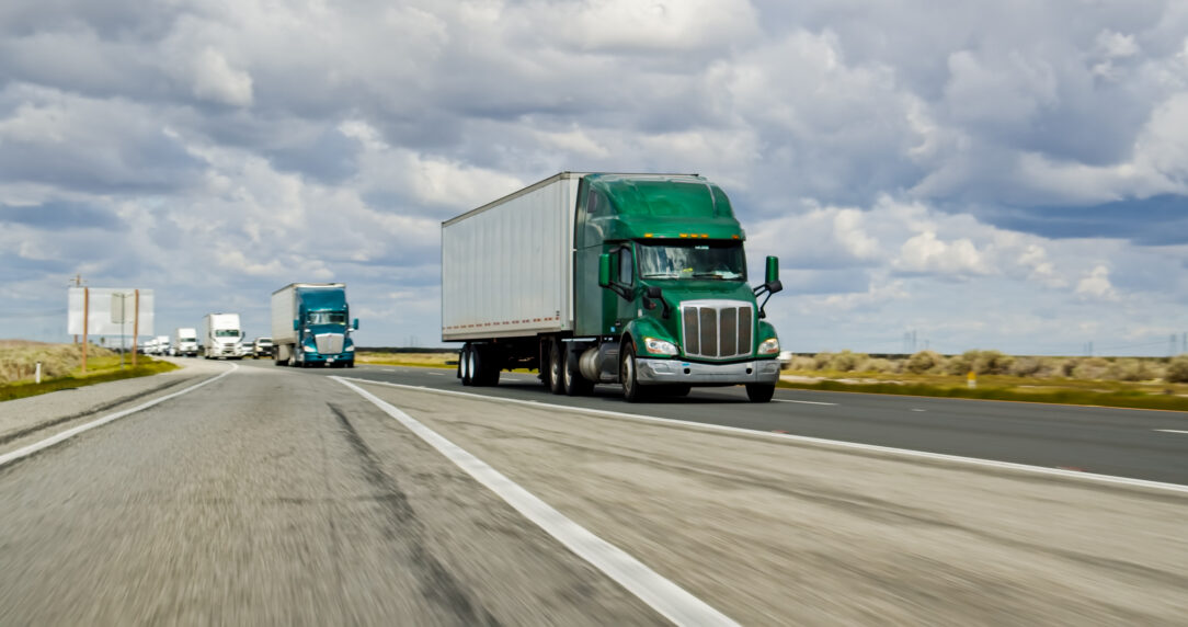 Green semi-truck driving on highway with multiple trucks in background under cloudy sky, motion blur suggesting speed.