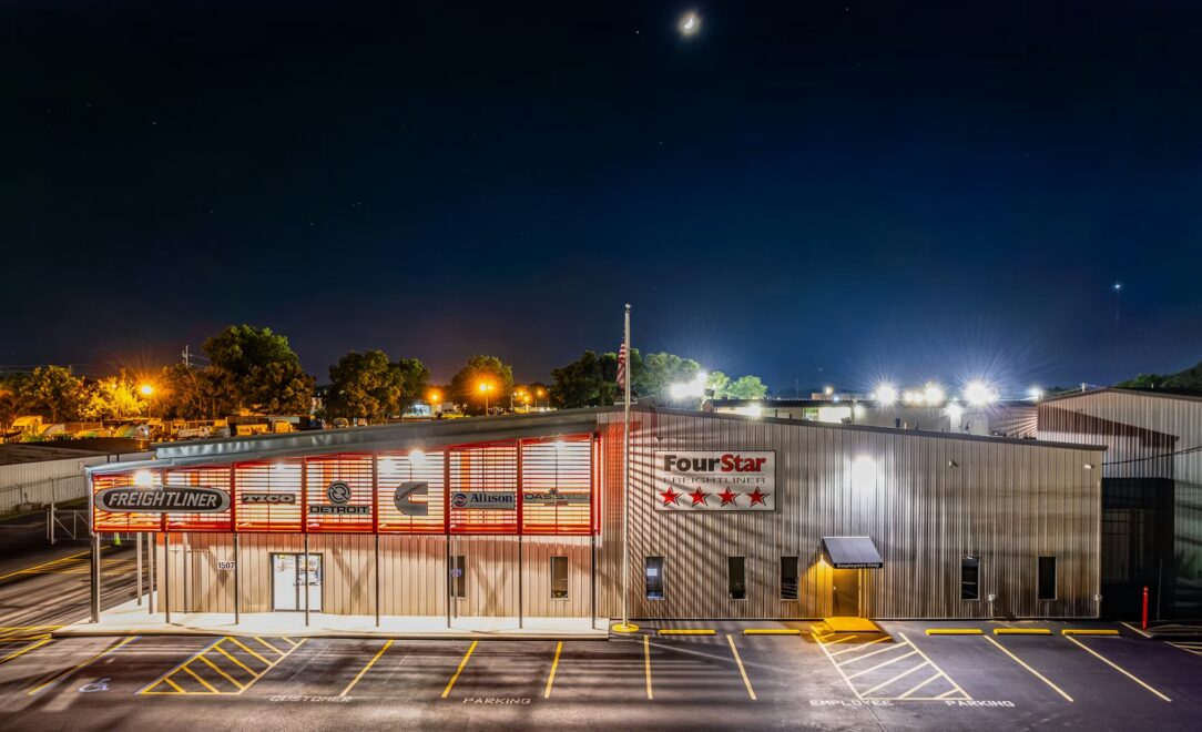 Nighttime view of a Freightliner building with illuminated red signage and a row of trees in the background, US flag on a flagpole in front.
