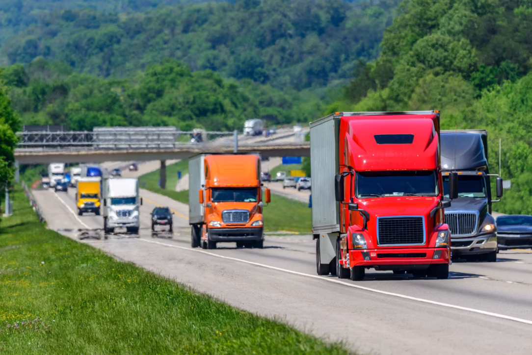 Line of colorful semi-trucks driving on a rural highway with green hills in the background.