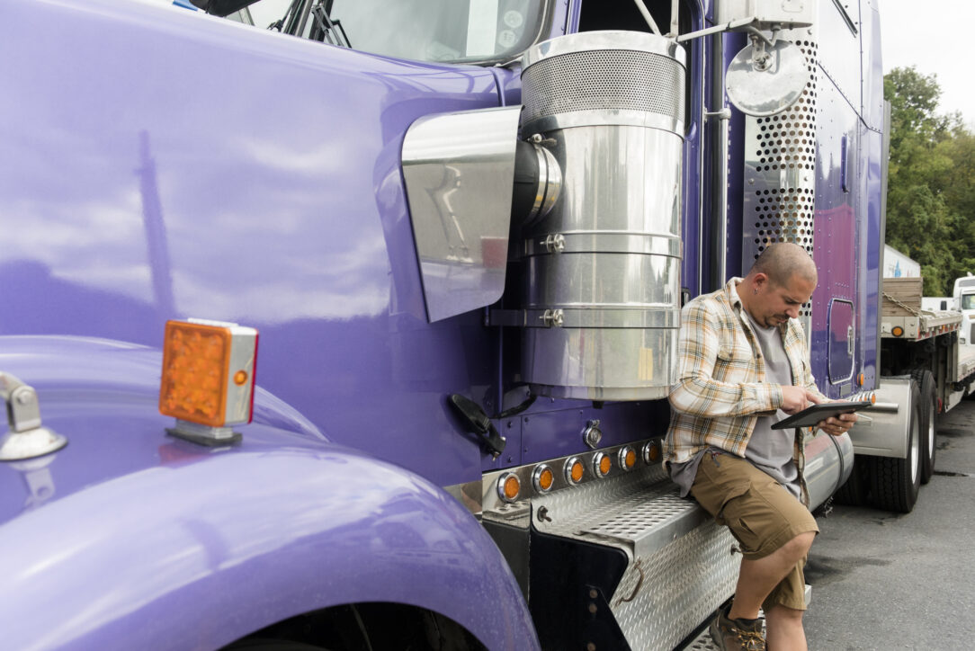 Man in a plaid shirt leans on the steps of a purple semi-truck cab, looking at a tablet in front of him.