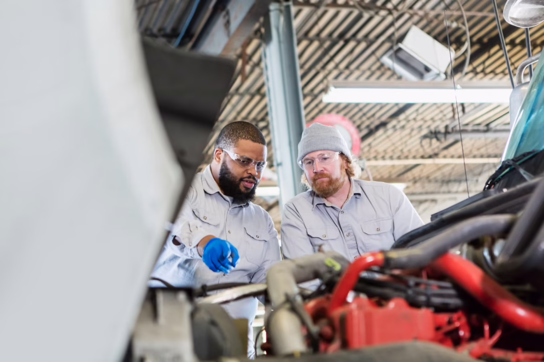 Two mechanics wearing safety glasses inspect a car engine in a bright workshop.